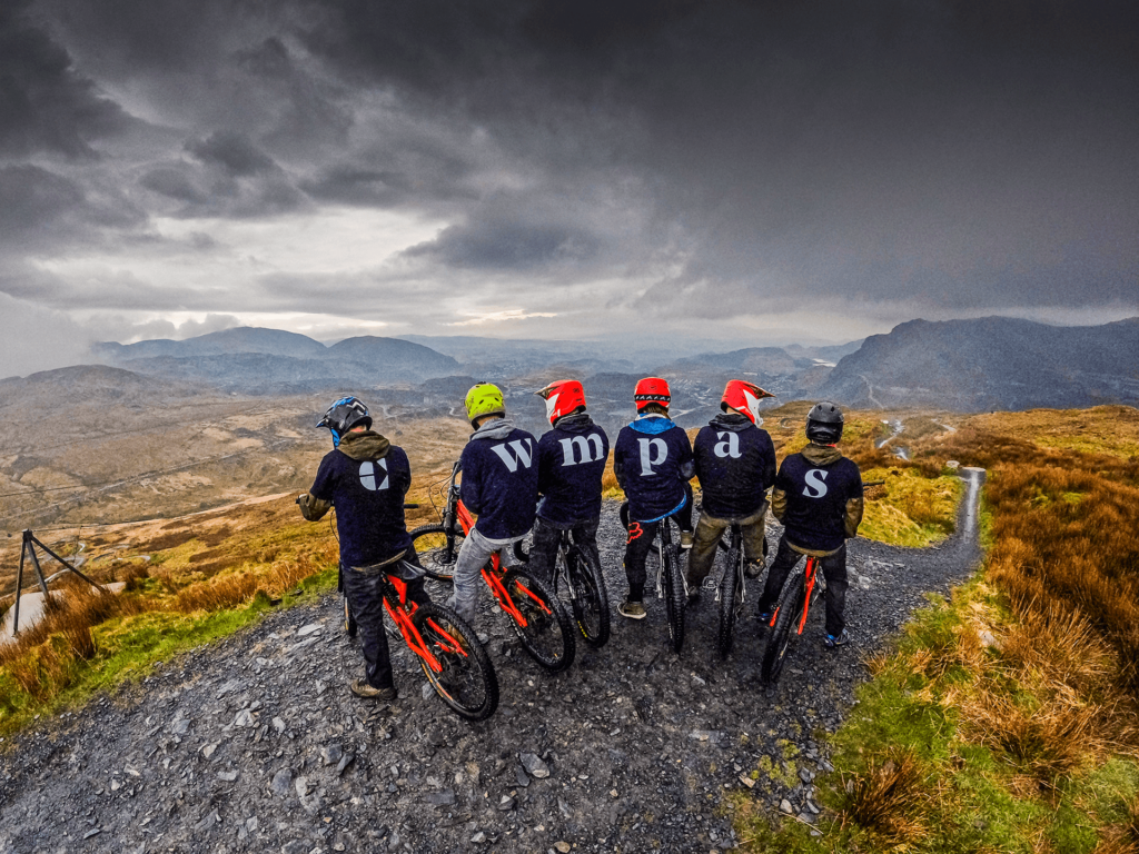 The Cwmpas Logo on the backs of t shirts of 6 bikers overlooking a valley.